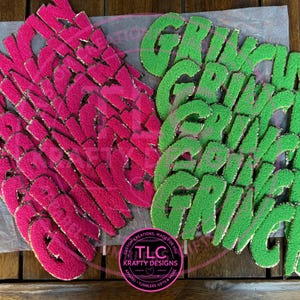 May include: A collection of pink and green fabric letters spelling "GRINCH". The letters have a textured, fuzzy appearance with gold trim. The image also includes a logo with the text "TLC Krafty Designs".