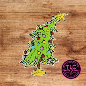 May include: A whimsical, green Christmas tree design with colorful ornaments and a yellow star topper. The tree has a yellow base and is set against a wood grain background. The design includes the text "TLC Krafty Designs".