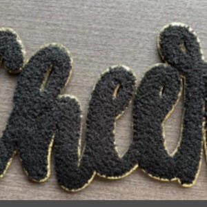 May include: A black, textured "Cheer" applique with gold trim. The letters are in a cursive font and are raised, giving a 3D effect. The applique is likely for clothing or accessories.
