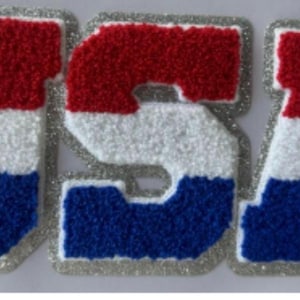 May include: A patriotic patch spelling out "USA" in red, white, and blue. The letters have a textured, felt-like appearance with a silver glitter border. The design is reminiscent of a varsity letter.