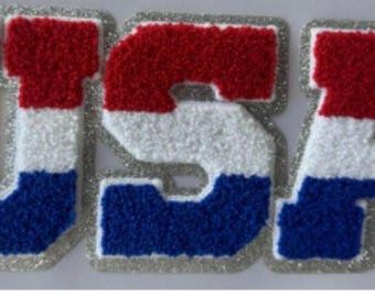 May include: A patriotic patch spelling out "USA" in red, white, and blue. The letters have a textured, felt-like appearance with a silver glitter border. The design is reminiscent of a varsity letter.