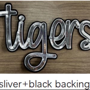 May include: A script-style patch featuring the word tigers in silver sequins with a black border and backing. The letters are connected in a cursive font, creating a single decorative piece with a reflective, metallic finish on the surface.