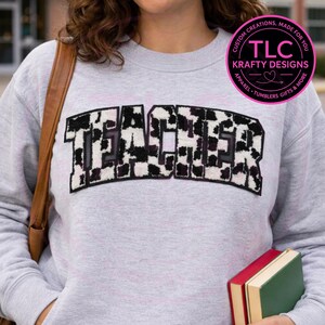 May include: A light gray sweatshirt with the word "TEACHER" in large, black and white cow print letters across the chest. The sweatshirt also has a small, circular logo with text that reads "TLC Krafty Designs".