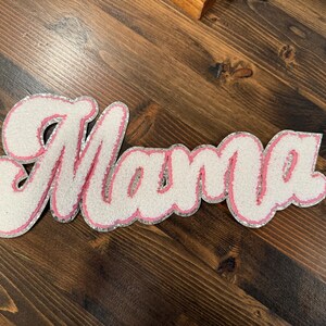 May include: A decorative sign spelling "Mama" in a cursive font. The letters are white with a pink outline and silver glitter trim. The sign is placed on a dark brown wooden surface.