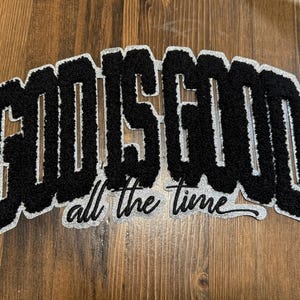 May include: Black and white patch with the text "GOD IS GOOD" in a varsity-style font, with the words "all the time" below. The patch has a textured, fuzzy appearance and a silver border.