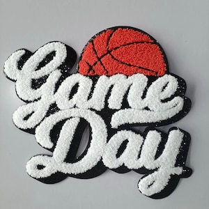 May include: A decorative patch with the words "Game Day" in white, textured lettering outlined in black. A red basketball with black lines is above the word "Day". The patch has a glittery finish.