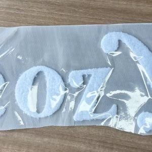 May include: Light blue felt letters spelling "COZY" are visible through a clear plastic sheet. The letters are in a cursive font and appear to be made of a soft, fuzzy material. The letters are on a light gray mesh background.