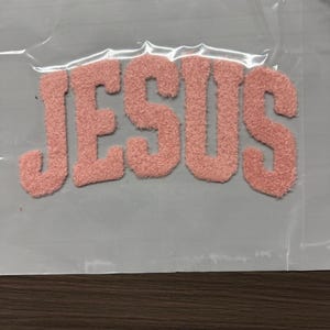 May include: A pink, fuzzy patch with the word "JESUS" in a bold, rounded font. The letters are a soft, textured material, possibly chenille or a similar fabric. The patch is on a light gray background.