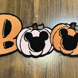 May include: A Halloween-themed patch spelling "Boo" with an orange letter and two pumpkin-shaped designs. The pumpkins have black Mickey Mouse silhouettes and are in shades of orange and pink. The patch has a black border.