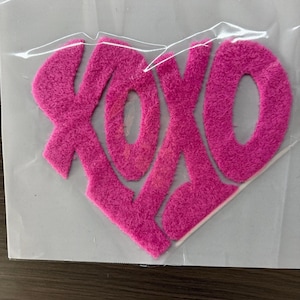 May include: A pink, heart-shaped felt applique with the letters "XOXO" in a textured, fuzzy font. The applique is packaged in clear plastic, ready for crafting or decoration.
