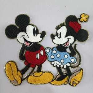 May include: Embroidered patch featuring Mickey Mouse and Minnie Mouse. Mickey is wearing red shorts and yellow shoes. Minnie is wearing a blue polka dot dress, red bow, and yellow shoes. Both characters have black fur and white faces.