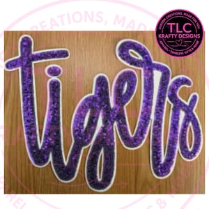 May include: A purple glitter script cutout featuring the word tigers with a white border. The letters have a dark purple to light purple gradient effect. A circular logo in the top right corner contains the text TLC KRAFTY DESIGNS.