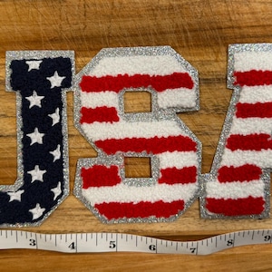May include: The word "USA" spelled out in individual fabric letters. The "U" is navy blue with white stars, the "S" and "A" are red and white striped. All letters have a silver glitter border. The letters are on a wooden surface.