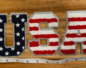 May include: The word "USA" spelled out in individual fabric letters. The "U" is navy blue with white stars, the "S" and "A" are red and white striped. All letters have a silver glitter border. The letters are on a wooden surface.