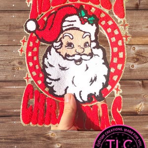 May include: A festive Christmas decoration featuring a smiling Santa Claus in a red hat and suit, surrounded by the words "MERRY CHRISTMAS" in red, glittery letters. The design includes a circular frame with gold accents and a small logo.