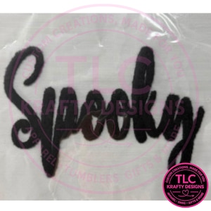 May include: A black iron-on transfer patch featuring the word Spooky written in a cursive script font. The letters are thick and textured, appearing as a dark silhouette against a light, semi-transparent backing material with a circular logo.