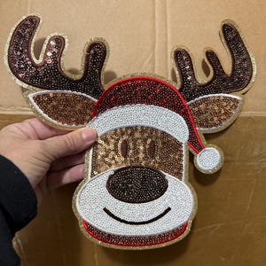 May include: A decorative Christmas reindeer head with antlers, a Santa hat, and a smiling face. The reindeer is embellished with sequins in shades of brown, gold, red, and white. The antlers and hat trim have a gold glitter border.