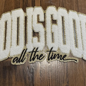 May include: White felt sign with the words "GOD IS GOOD" in large, stylized letters with gold glitter trim. Below, the phrase "all the time" is written in a black script font. The sign is placed on a wooden surface.