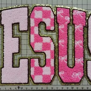 May include: A pink and gold embroidered patch spelling out the word "JESUS". The letters are in a varsity style, with a gold glitter border. The "E" has a pink and white checkered pattern, and the "J" and "S" have white bow accents.