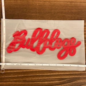 May include: A red felt applique in the shape of the word "Bulldogs" is displayed inside a clear plastic bag. The applique measures approximately 10 inches in length.