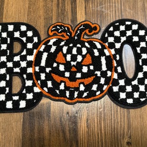 May include: A Halloween-themed decorative piece spelling "BOO" with a pumpkin in the center. The letters and pumpkin are black and white checkered, with an orange pumpkin face. The piece is placed on a wooden surface.