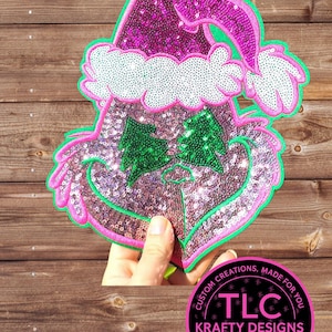 May include: A sequined Grinch face patch with a Santa hat. The Grinch's face is silver with green sequined eyes and a pink outline. The Santa hat is pink with a white trim and green outline. The patch is held in front of a wooden background.