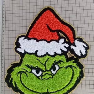 May include: A green Grinch face patch with a red Santa hat and white trim. The Grinch has a black outline, a gold border, and a textured appearance. The patch is on a grid background.