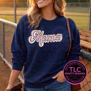 May include: Navy blue sweatshirt with the word "Mama" in a baseball-themed design. The word "Mama" is embroidered with red stitching and a white outline, evoking a baseball. The sweatshirt also features the text "TLC Krafty Designs" in a circular logo.
