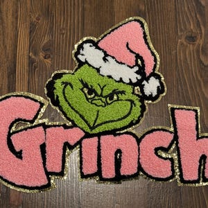 May include: A green Grinch face wearing a pink Santa hat with white trim, above the pink word "Grinch". The letters and Grinch face have a gold border. The design is on a wooden surface.