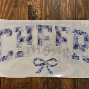 May include: A white fabric patch with the words "CHFFD Mom" in blue and silver rhinestones. A blue bow is at the bottom. The patch is on a wooden surface.