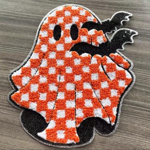 May include: A Halloween-themed rug in the shape of a ghost with a checkered orange and white pattern. The ghost has black eyes and mouth, with two black bat shapes. The rug is outlined with a silver beaded trim.