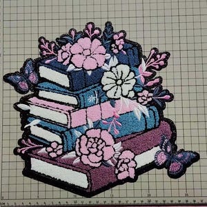 May include: Embroidered patch featuring a stack of books in shades of blue, pink, and purple, adorned with pink and white flowers and two butterflies. The patch has a black glitter border.