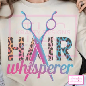 May include: A cream-colored sweatshirt featuring a graphic of crossed hair shears with a pink and blue gradient. The word HAIR is printed in large block letters with a leopard print pattern, and the word whisperer is in pink script below.