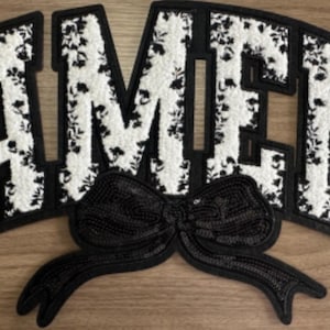 May include: A black and white patch spelling "AMEN" with a black bow. The letters are white with a floral pattern and outlined in black. The bow is made of black sequins. The patch is on a wooden surface.