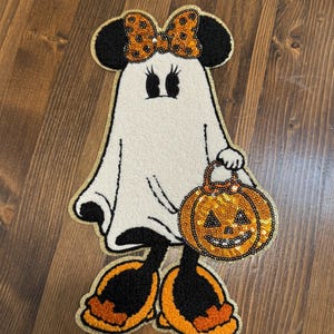 May include: A Halloween-themed decorative item featuring a ghost character with mouse ears and a bow. The ghost is holding a sequined pumpkin. The design has black, orange, and gold accents, with a gold border.