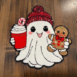 May include: A whimsical, white ghost-shaped rug with a red sequined beanie, holding a red cup of whipped cream and a gingerbread man. The rug has a black border and is set against a wooden background.