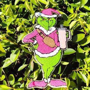 May include: A green cartoon character wearing a pink Santa hat and sweater, holding a mug. The character has a grumpy expression and is set against a backdrop of green leaves. The character is wearing a brown satchel.