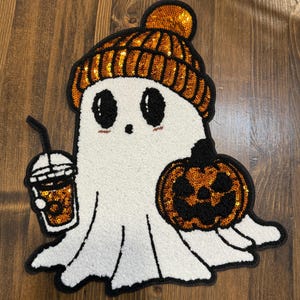 May include: A white ghost-shaped Halloween decoration with a black outline. The ghost wears an orange sequined beanie and holds a pumpkin and a coffee drink, both with orange sequins. The ghost has black eyes and a small smile.