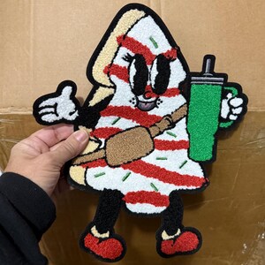 May include: A tufted textile patch shaped like a slice of cake with red and white stripes, green sprinkles, and a cartoon face. The character wears a brown crossbody bag, holds a green tumbler with a straw, and has black legs with red shoes.