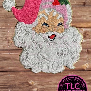 May include: A festive Santa Claus design with a pink hat, white beard, and rosy cheeks. The Santa Claus has blue eyes and a smiling red mouth. The design is outlined in gold and set against a wooden background. The image includes a circular logo with text.