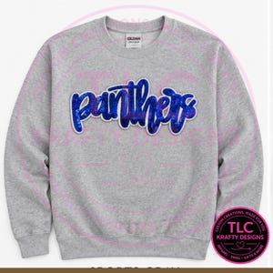 May include: A light gray crewneck sweatshirt featuring the word panthers in a blue glitter script font across the chest. The text has a white outline and a subtle drop shadow effect. The garment has long sleeves and a ribbed collar and cuffs.