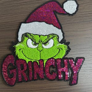 May include: A green Grinch face with an angry expression, wearing a red sequined Santa hat with white trim. The word "GRINCHY" is spelled out in red sequins below the face. The design is on a brown background.