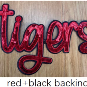 May include: A script-style iron-on patch featuring the word tigers in bright red sequins. The lettering is outlined with a dark black border, providing a high-contrast look. The entire design is set against a black backing material.