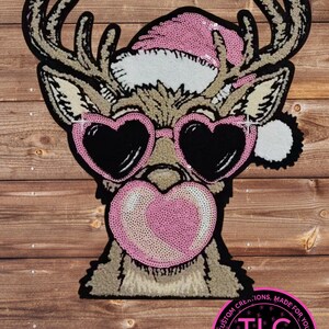 May include: A whimsical patch featuring a reindeer with heart-shaped sunglasses and a pink Santa hat. The reindeer is blowing a heart-shaped bubblegum bubble. The patch has a black border and the text "TLC Krafty Designs".