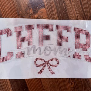 May include: A white fabric sheet with the words "CHEER mom" in red and silver rhinestones. A red rhinestone bow is at the bottom. The text is in a stylized font, perfect for cheerleading apparel.