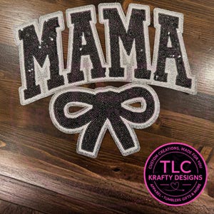 May include: A decorative patch set featuring the word "MAMA" in large, sequined letters with a silver border. Below, a sequined bow with a silver border. The patches are on a wooden surface.