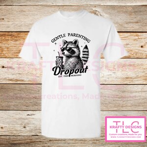 May include: White t-shirt featuring a black and white raccoon wearing sunglasses and holding a leopard print cup. The text reads "GENTLE PARENTING Dropout EST. THIS MORNING". The shirt has a crew neck and short sleeves.