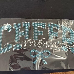 May include: Black t-shirt with a rhinestone design. The design says "CHEER mom" in a script font, with a blue bow at the bottom. The letters are made of blue and clear rhinestones.