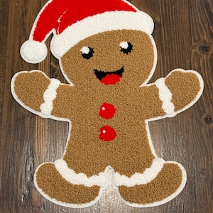 May include: A brown gingerbread man rug with a Santa hat. The rug has a white border and white accents on the arms and legs. The gingerbread man has a smiling face with red buttons and a red mouth. The rug is on a wooden surface.
