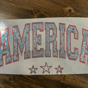 May include: A white rectangular patch with the word "AMERICA" in large, sparkling letters. The letters are outlined in red and filled with blue and clear rhinestones. Three stars are below the word.